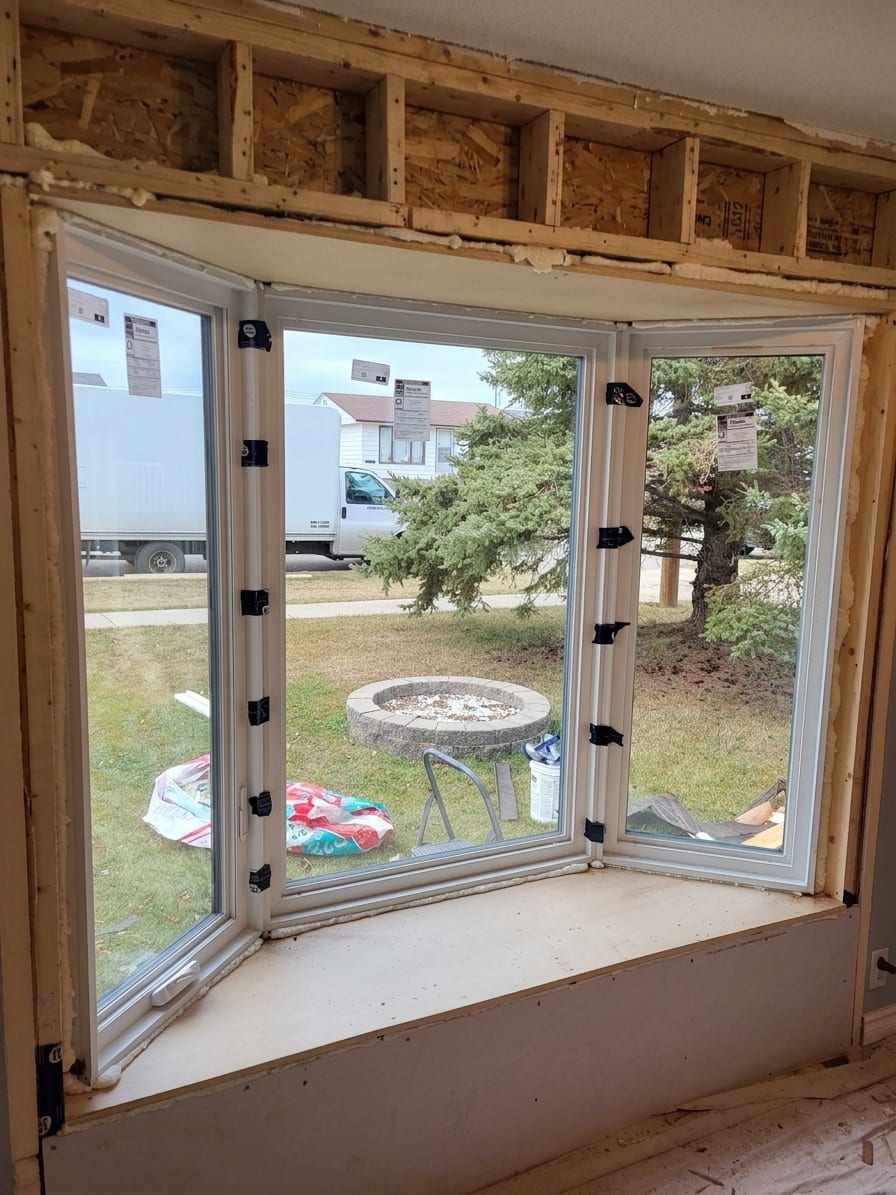 A bay window part-way through installation, seen from inside: three white-framed units with shipping labels and transit clips still attached, set into an opening with exposed studs, sheathing and spray foam above.