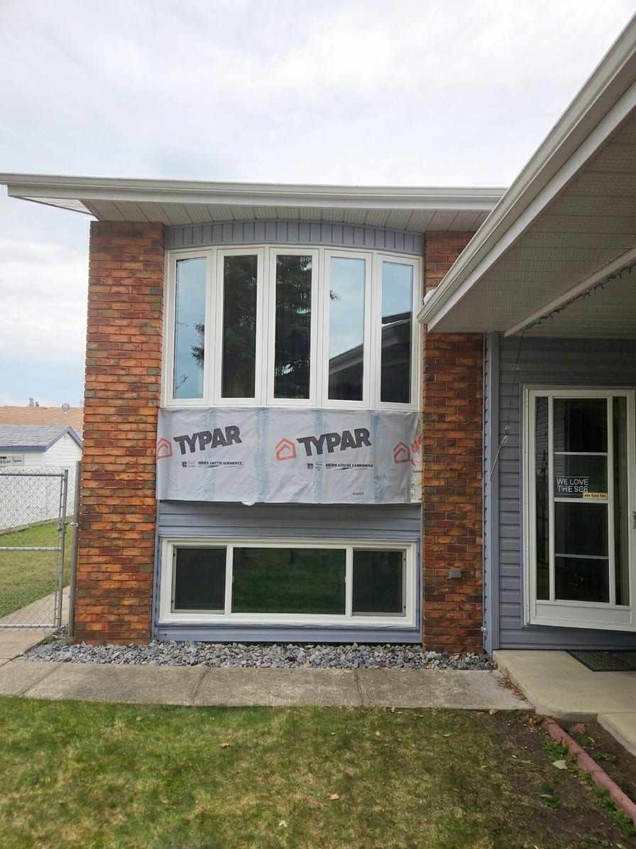 The front of a bi-level house between two brick columns, with a five-panel white bow window on the upper floor, a three-panel window at basement level, and house wrap exposed on the wall between them.