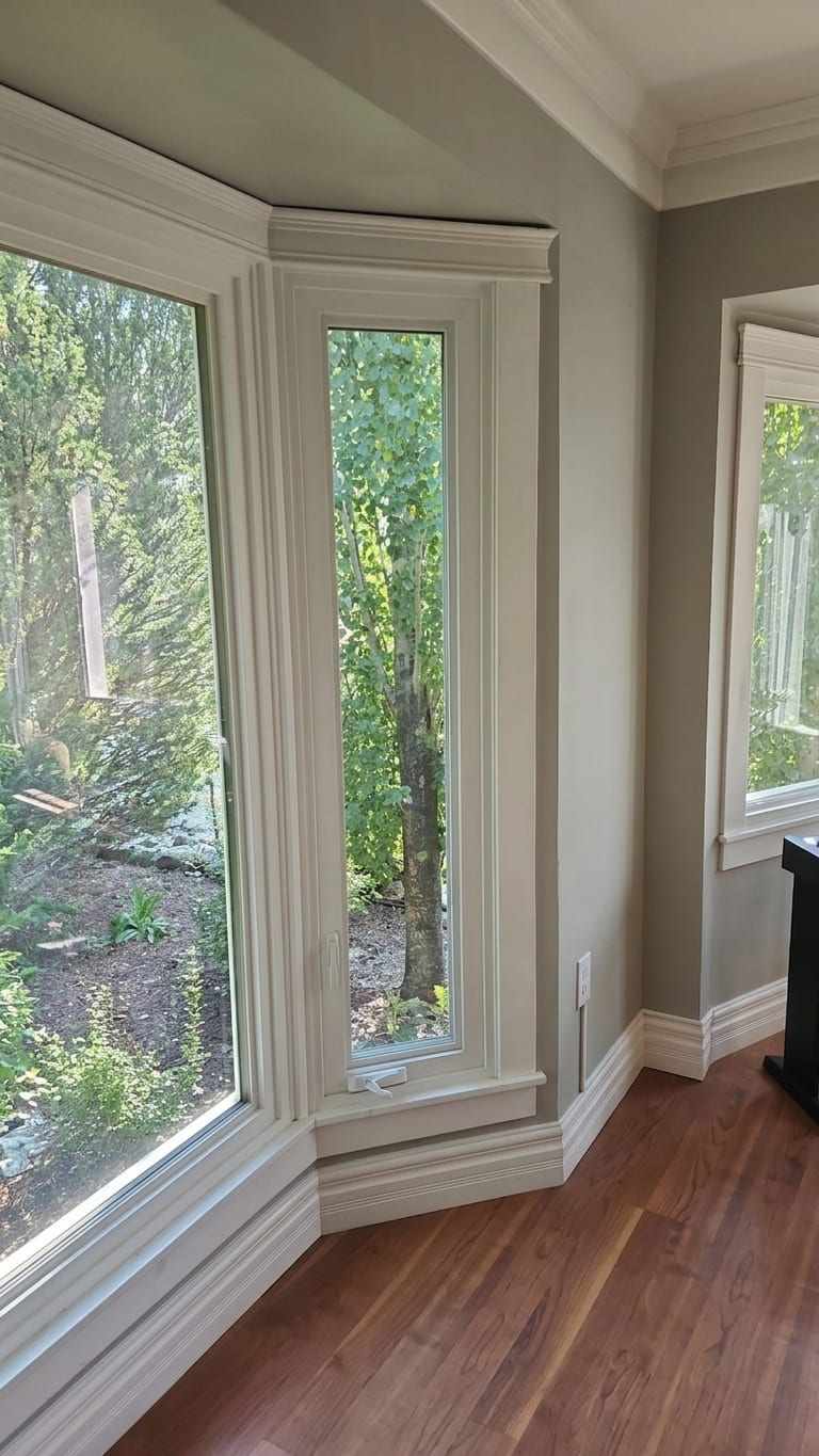 An interior corner with a tall narrow casement window showing its folding crank handle and lock, beside a large fixed picture window, both in painted timber casings above a hardwood floor.