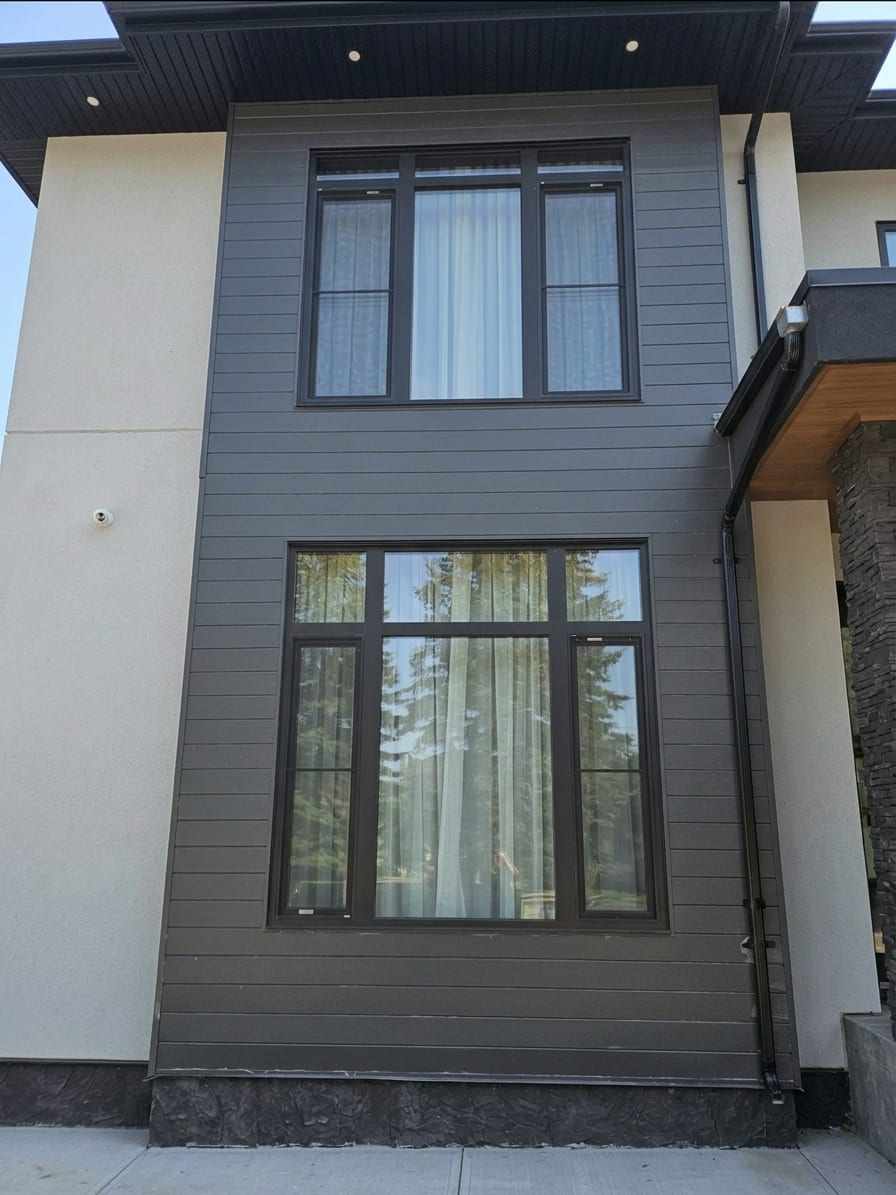 A two-storey elevation clad in dark horizontal siding and pale stucco, with two window groups stacked one above the other, each a wide fixed pane flanked by narrower operating units under a row of transom lights.